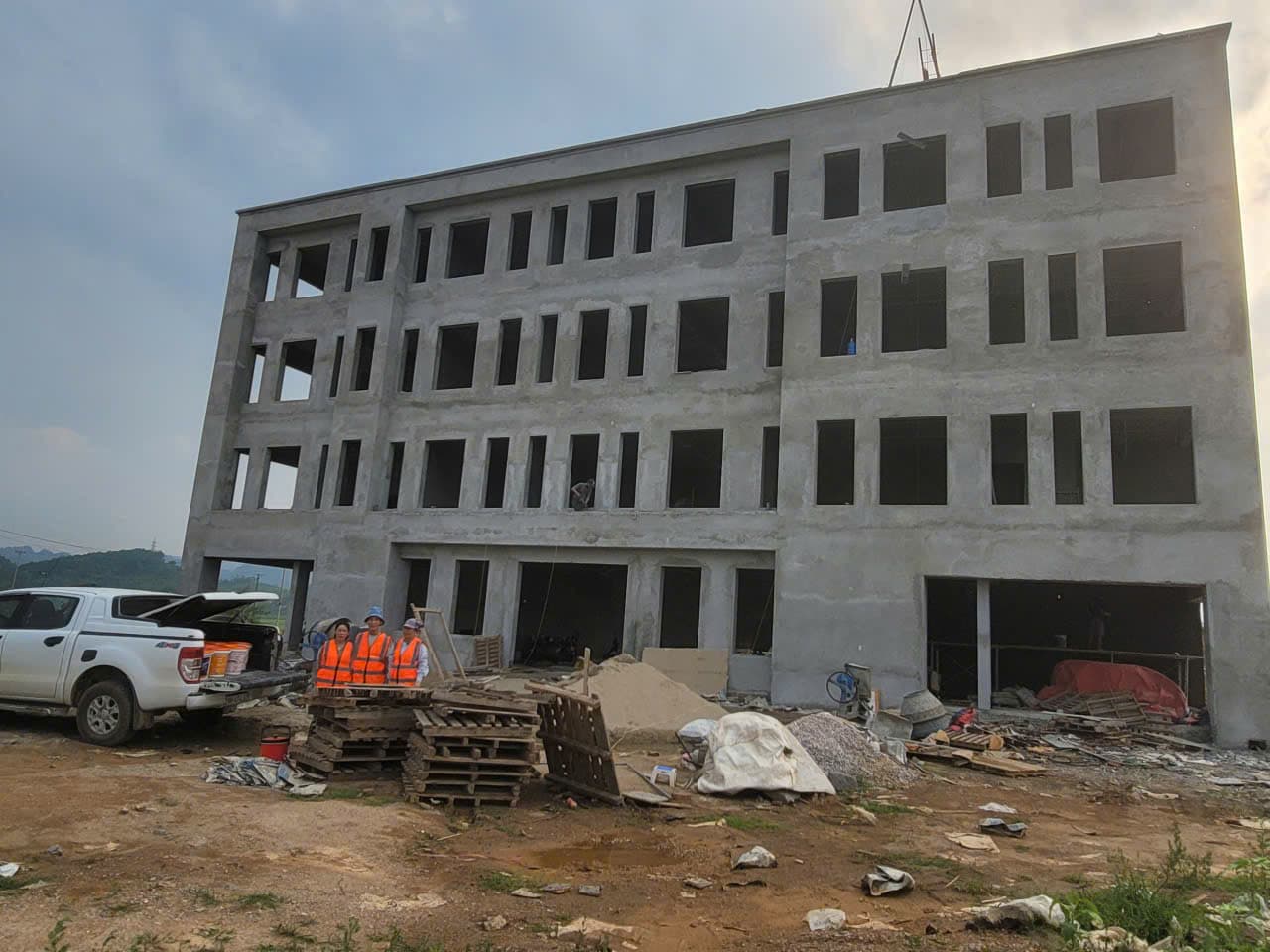 Công trình bê tông bốn tầng đang thi công, thợ mặc áo phản quang đứng cạnh xe bán tải và vật tư.
