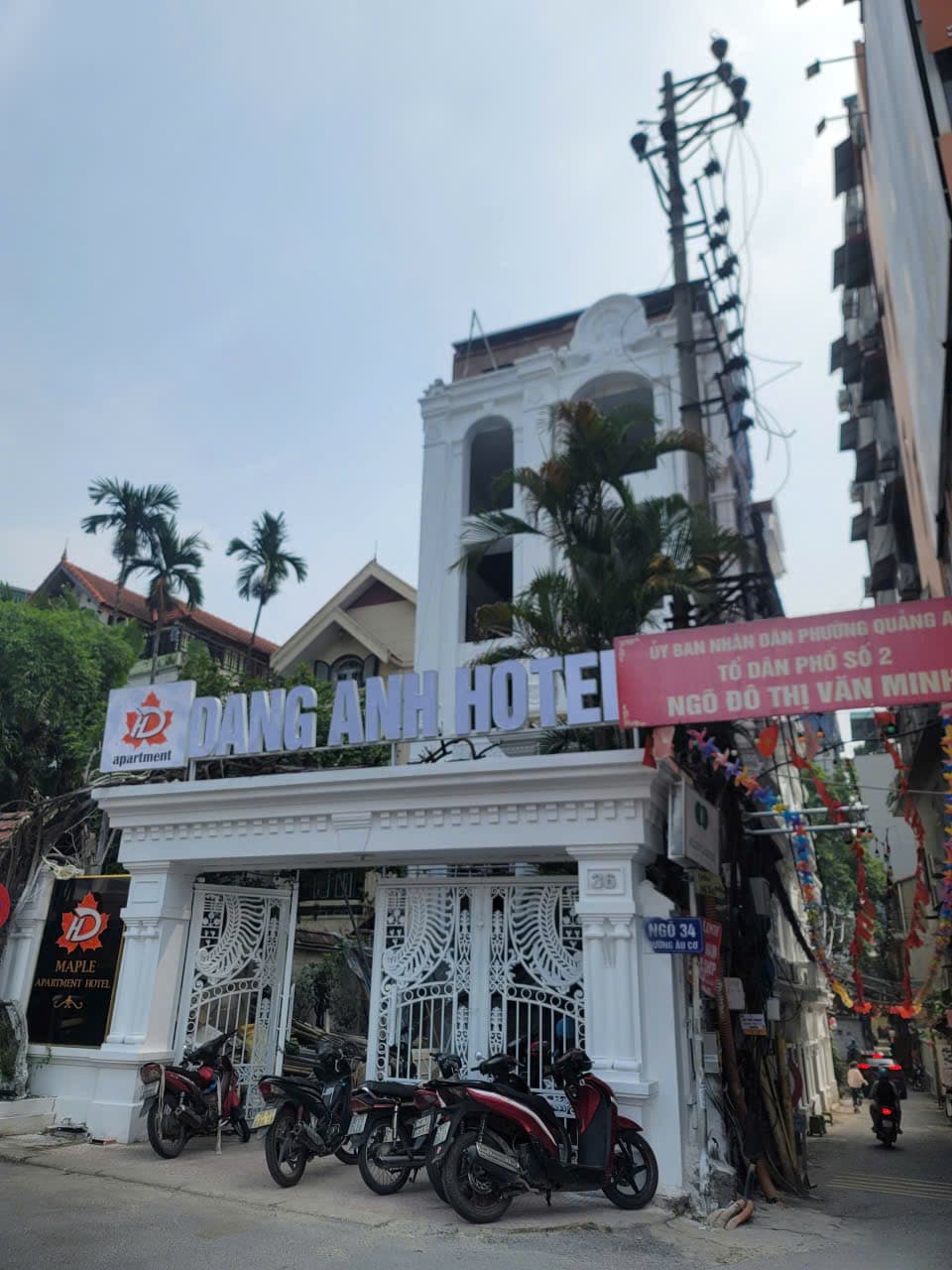 Tòa nhà trắng mặt phố với cổng sắt lớn, bảng hiệu và dãy xe máy đậu kín vỉa hè.