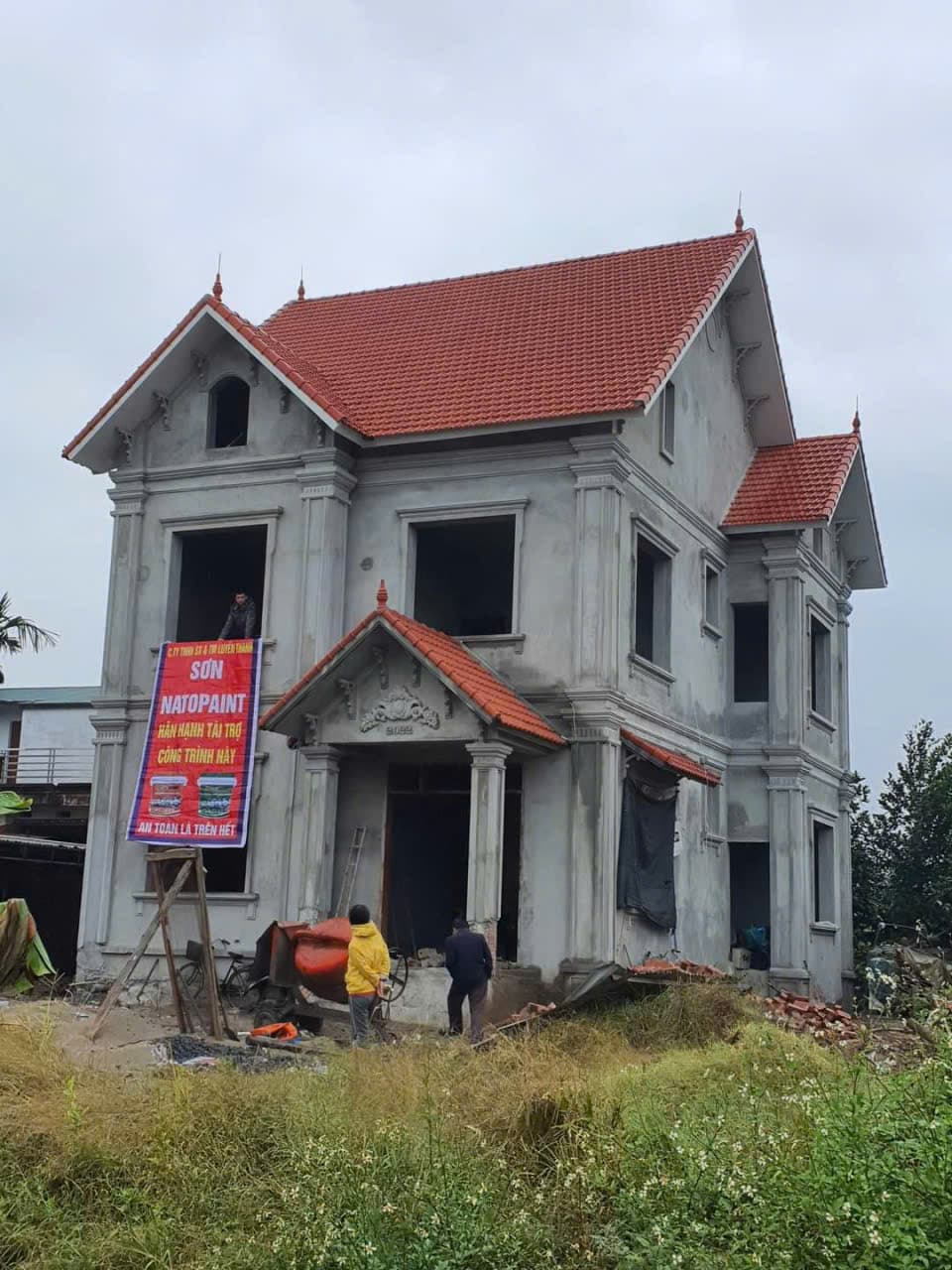 Nhà hai tầng mái ngói đỏ đang xây, băng rôn treo trước cửa và thợ làm việc bên dưới.