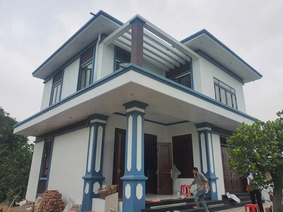 Nhà hai tầng cột xanh trắng có giàn lam trên sân thượng, gạch và vật tư còn để trước hiên.