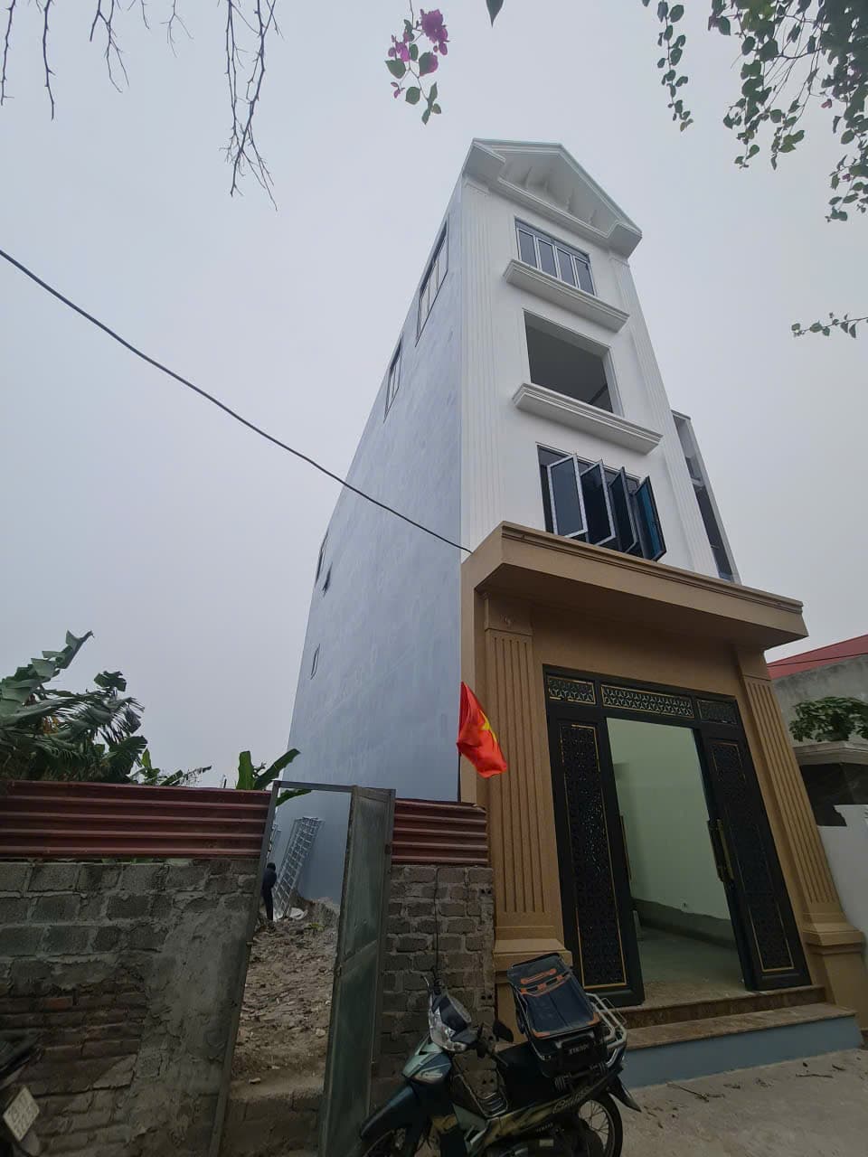 Nhà phố bốn tầng mặt tiền hẹp, tường trên trắng dưới xanh, cổng sắt trang trí và xe máy trước cửa.